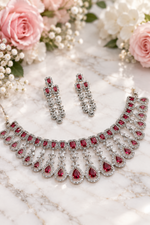 Load image into Gallery viewer, A silver-toned jewelry set featuring a statement necklace and matching drop earrings, embellished with red gemstones and clear crystals, styled on a textured cream fabric with pink flowers in the background.